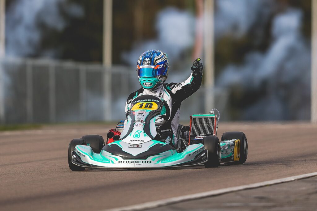 Driver in a teal, white and black Rosberg Racing Academy kart, number 10, one hand raised